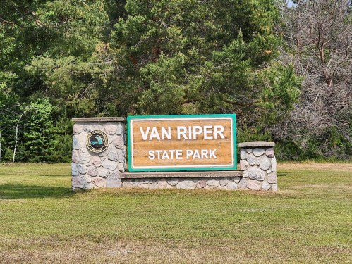 Sign for Van Riper State Park, featuring a wooden plaque with stone supports, surrounded by greenery.