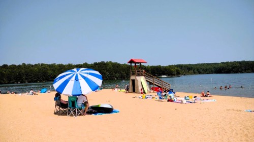 A sandy beach with people relaxing under umbrellas, a lifeguard tower, and a calm lake in the background.