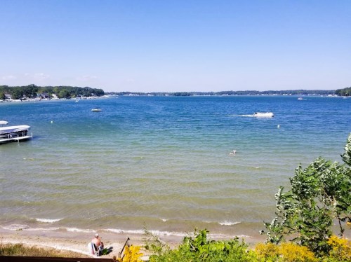 A serene lake view with boats, a sandy shore, and clear blue skies, surrounded by greenery.