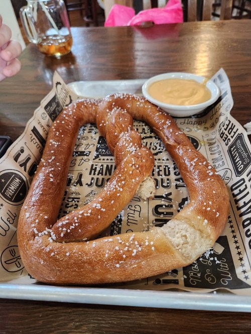 A large, twisted pretzel sprinkled with salt, served on a tray with a small bowl of cheese dip.