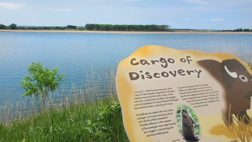 A sign titled "Cargo of Discovery" near a river, with greenery and a clear blue sky in the background.