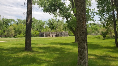 A rustic wooden structure surrounded by lush green trees and grass under a partly cloudy sky.
