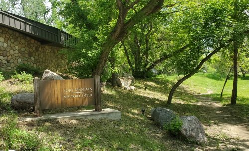 Sign for Fort Mandan Visitor Center surrounded by trees and rocks, with a grassy area in the background.
