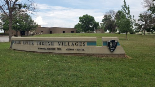 Sign for the Knife River Indian Villages National Historic Site and Visitor Center, surrounded by green grass and trees.