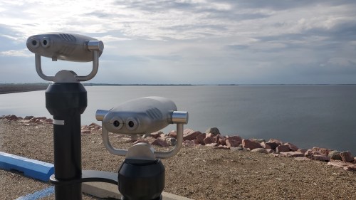 Two binoculars on a stand overlooking a calm body of water, with a cloudy sky in the background.