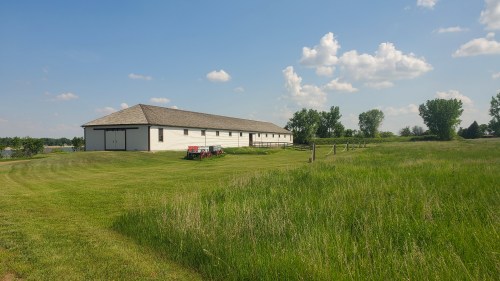 A large white barn sits beside a grassy field under a blue sky with fluffy clouds. Trees line the background.