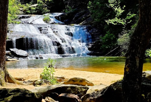 A serene waterfall cascades into a calm pool, surrounded by lush greenery and sandy shores.