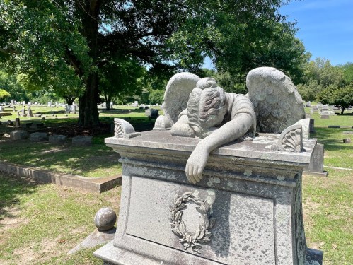 A stone angel statue rests on a grave, with its head bowed and arms draped over a tomb in a serene cemetery setting.