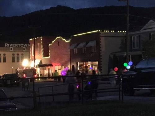 A nighttime scene of a small town with festive lights and people walking, some holding colorful balloons.