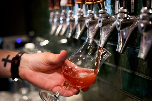 A hand pours a pink drink from a tap into a glass at a bar with green tiled walls.