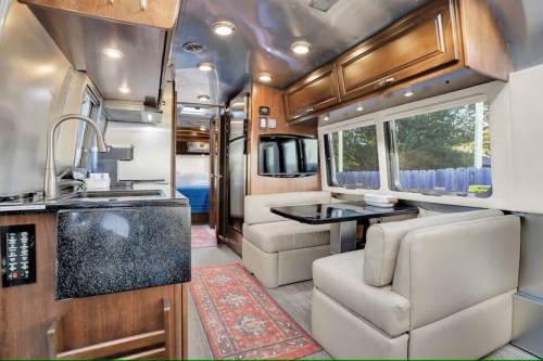 Interior of a modern RV featuring a kitchen area, dining table, and seating with large windows and wooden cabinetry.