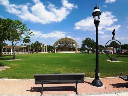 A park scene featuring a stage, a bench, a lamp post, and a sculpture, with blue skies and scattered clouds.