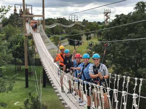 A group of children in helmets smiles while crossing a rope bridge in an outdoor adventure park.