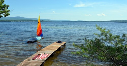 A colorful sailboat is docked at a wooden pier on a calm lake, surrounded by trees and mountains under a clear blue sky.