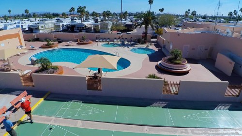 Aerial view of a pool area with palm trees, lounge chairs, and a shuffleboard court in a sunny resort setting.