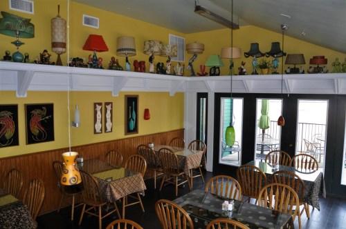 A cozy restaurant interior with wooden tables, colorful lamps on shelves, and large windows letting in natural light.