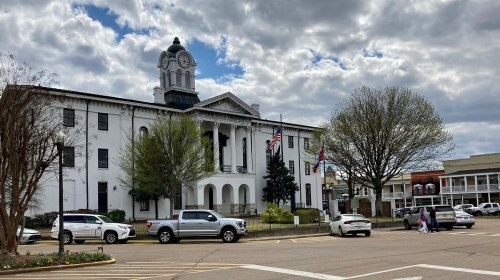 Historic white courthouse with a clock tower, surrounded by trees and cars, under a cloudy sky.
