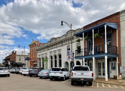 Historic buildings line a street with parked cars, under a partly cloudy sky.