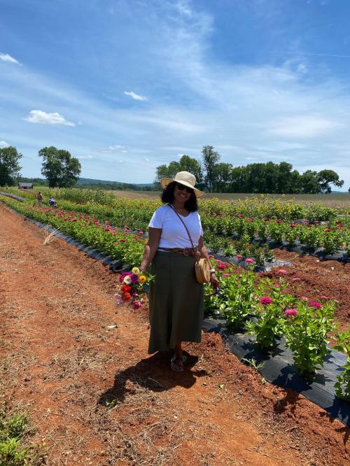 A woman in a white shirt and green skirt stands in a flower field, holding a bouquet, with blue skies above.