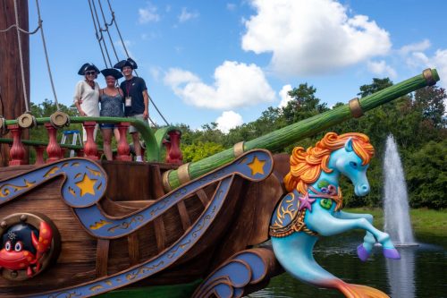 Two people in pirate hats pose on a colorful ship with a whimsical horse figure, surrounded by greenery and a fountain.