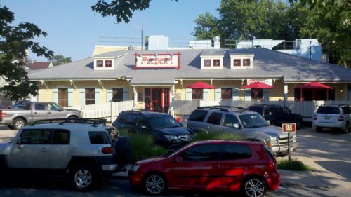 A yellow restaurant with a red sign, surrounded by parked cars and trees, featuring outdoor seating under red umbrellas.