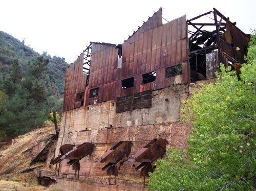 A weathered, abandoned building with rusted metal walls and a crumbling foundation, surrounded by trees and hills.