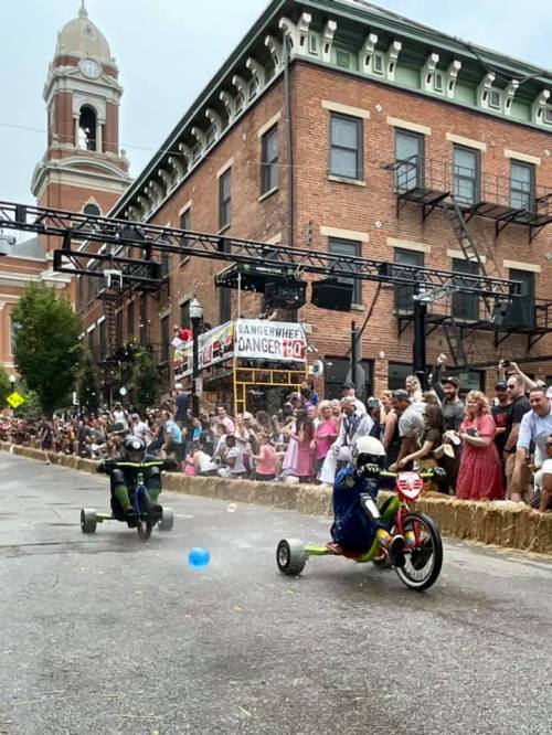 Two children racing on tricycles down a street lined with spectators, with a festive atmosphere and buildings in the background.