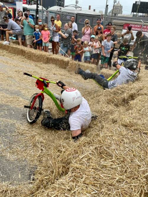A child falls off a small bike during a race on a straw-covered street, while a crowd watches and cheers nearby.