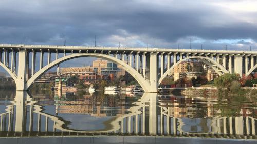 A bridge with arches reflected in calm water, surrounded by buildings and a cloudy sky.