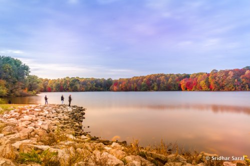 A serene lake surrounded by colorful autumn trees, with silhouettes of people walking along the rocky shore.