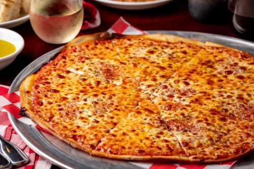 A close-up of a cheese pizza on a metal tray, with a glass of white wine in the background.