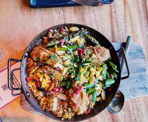 A vibrant dish featuring rice, grilled vegetables, pomegranate seeds, and herbs in a round metal serving pan.