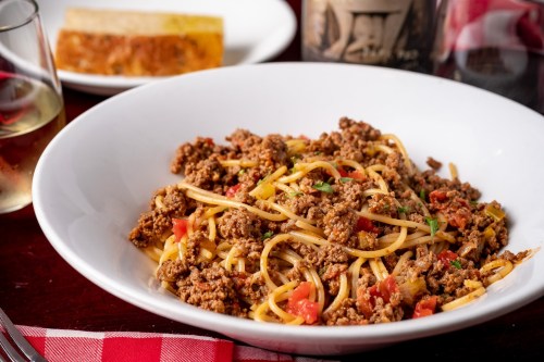 A bowl of spaghetti topped with seasoned ground meat and tomatoes, served with a slice of bread and a glass of wine.