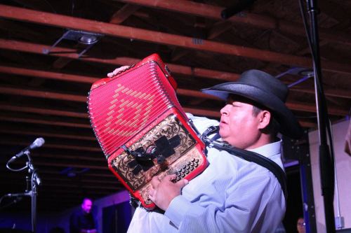 A musician in a cowboy hat passionately plays a red accordion on stage, with microphones in the background.