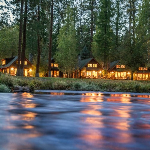 Cozy cabins with warm lights reflect on a calm river, surrounded by tall trees at dusk.