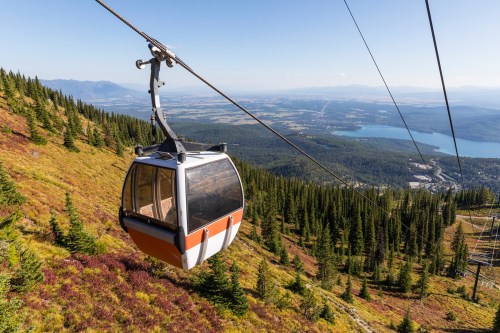 A gondola glides over a colorful mountain landscape with trees and a lake in the distance under a clear blue sky.