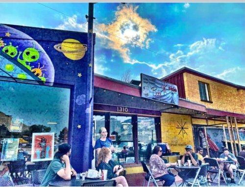 A colorful café scene with people sitting outside, featuring a mural of aliens and a bright sky with clouds.