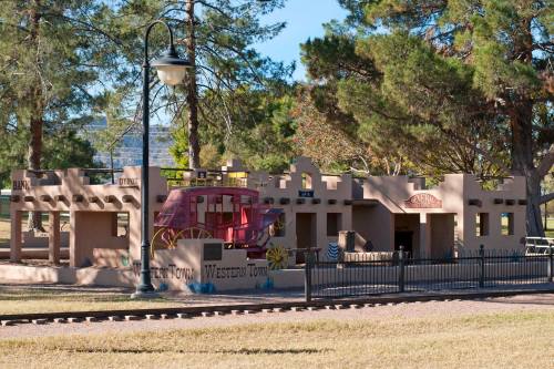 A colorful, western-themed play structure with a train and buildings, surrounded by trees and a grassy area.