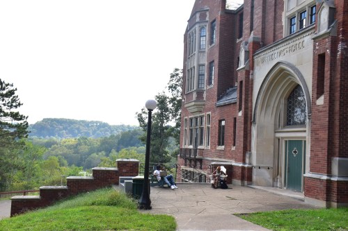A brick building with large windows overlooks a green landscape, with two people sitting on the steps.
