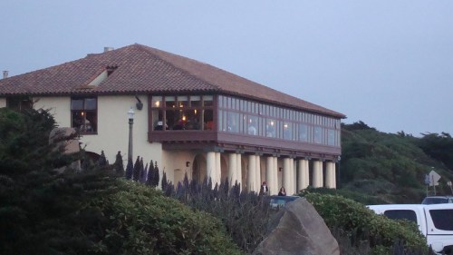 A coastal building with large windows, surrounded by greenery and a rocky landscape, during twilight.