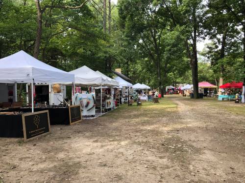 A tree-lined path with various vendor tents set up for a market, showcasing crafts and goods in a natural setting.