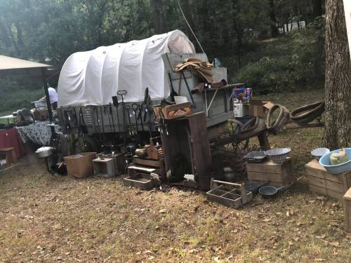 A vintage wagon covered with a white tarp, surrounded by various tools and containers in a wooded area.