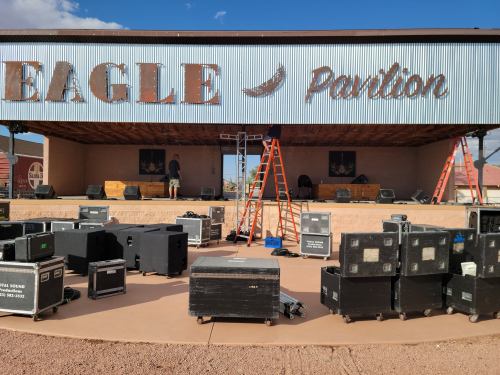A stage at Eagle Pavilion with sound equipment, ladders, and a person preparing for an event under a blue sky.