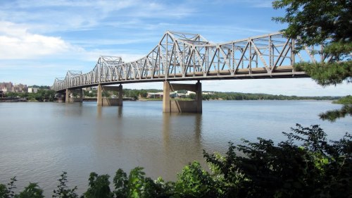 A metal bridge spans a calm river, with greenery in the foreground and a clear blue sky above.