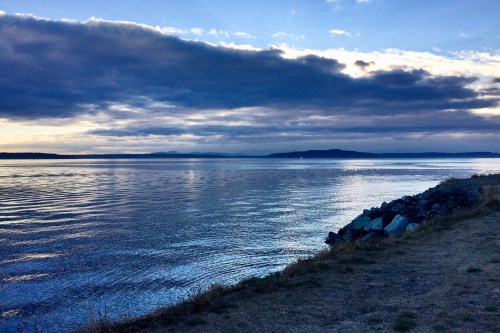 A serene waterfront scene at dusk, featuring calm waters and a cloudy sky reflecting soft blue hues.