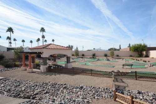 A mini-golf course with palm trees, a gazebo, and rocky landscaping under a blue sky with wispy clouds.