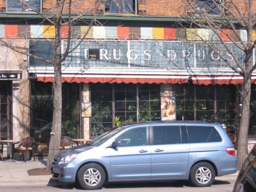 A blue minivan parked in front of a storefront with colorful awnings and large windows displaying plants inside.