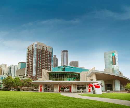 A modern building with a large glass entrance, surrounded by green grass and city skyscrapers under a blue sky.