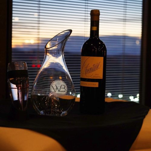 A wine decanter and bottle sit on a table with a glass of water, against a sunset backdrop and city lights.