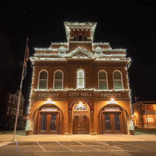 Historic brick building decorated with holiday lights, featuring "Fire Dept" and "City Hall" signs, at night.